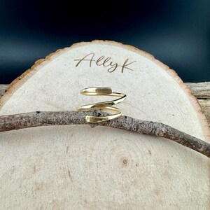 May include: A gold-colored spiral ring with a hammered texture. The ring is displayed on a small branch, set against a light wood slice with the name "Allyk" engraved on it. The background is dark.