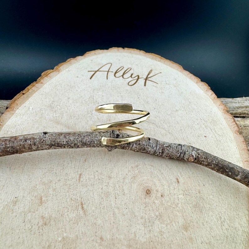 May include: A gold-colored spiral ring with a hammered texture. The ring is displayed on a small branch, set against a light wood slice with the name "Allyk" engraved on it. The background is dark.