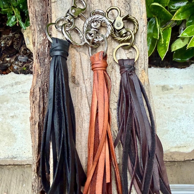 May include: Three leather tassel keychains in black, brown, and tan. Each keychain has a metal ring and clasp. The tassels are made of long, thin strips of leather. The keychains are displayed against a wooden background.