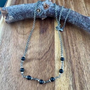 May include: A silver-toned necklace with black beads. The necklace features a delicate chain and is accented with small, round black beads. The beads are interspersed with silver-toned metal accents. The necklace is displayed on a wooden surface.