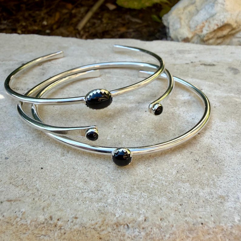 Op de afbeelding: Drie zilverkleurige armbanden met zwarte edelsteenaccenten. De armbanden hebben een eenvoudig, open ontwerp met een gladde, gepolijste afwerking. E&eacute;n armband heeft een ovaalvormige steen, de andere ronde stenen.