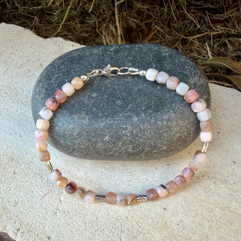 May include: A beaded bracelet featuring small, cube-shaped pink and white stones arranged in a continuous loop. The bracelet includes metallic silver-toned spacer beads and a silver-toned lobster claw clasp closure, creating a delicate accessory.