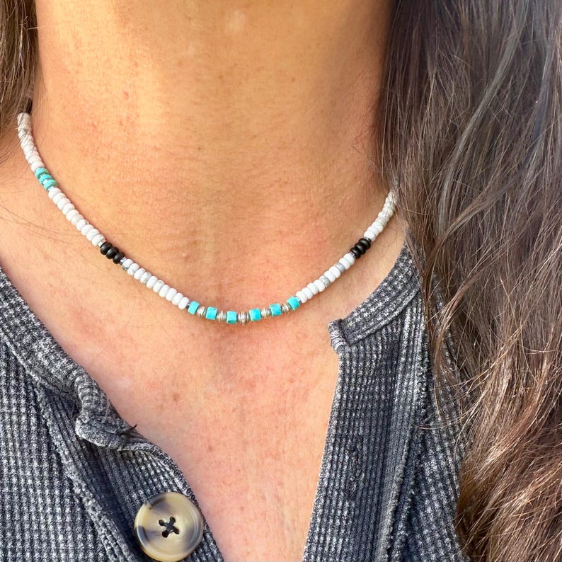 May include: A beaded necklace featuring white, black, and turquoise beads. The necklace has a delicate design with small, round white beads interspersed with turquoise and black beads. The necklace is worn around the neck.