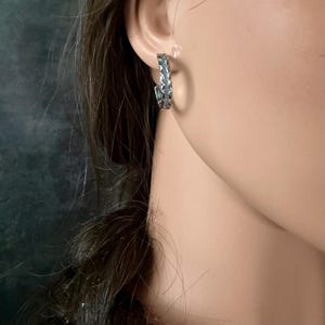 May include: A silver-toned hoop earring with a detailed, textured design. The earring is worn on the ear, showcasing its intricate pattern. The jewelry piece is a stylish accessory.