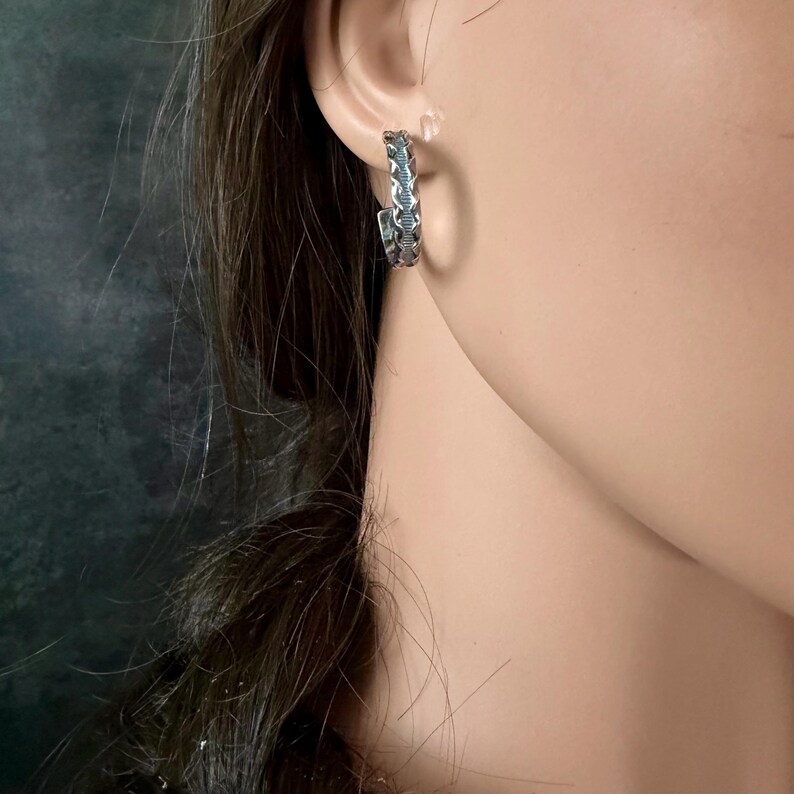 May include: A silver-toned hoop earring with a detailed, textured design. The earring is worn on the ear, showcasing its intricate pattern. The jewelry piece is a stylish accessory.