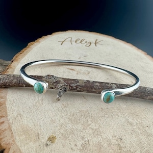 K&ouml;nnte beinhalten: Ein silberfarbenes Armreif-Armband mit zwei t&uuml;rkisfarbenen Steinen. Das Armband wird auf einer Holzoberfl&auml;che pr&auml;sentiert. Der Text "Allyk" ist im Hintergrund zu sehen.