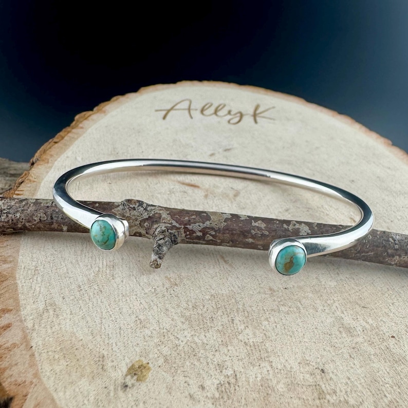 K&ouml;nnte beinhalten: Ein silberfarbenes Armreif-Armband mit zwei t&uuml;rkisfarbenen Steinen. Das Armband wird auf einer Holzoberfl&auml;che pr&auml;sentiert. Der Text "Allyk" ist im Hintergrund zu sehen.