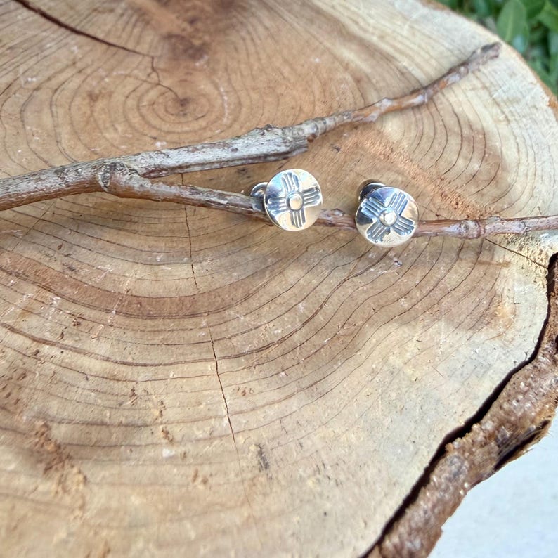 May include: A pair of round silver stud earrings featuring a stamped Zia sun symbol design. Each earring has a polished metallic finish with a central dot surrounded by four radiating lines, securely attached to a thin, natural wooden branch.