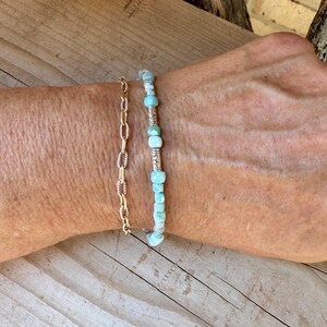 Puede incluir: Una pulsera de cadena dorada con eslabones ovalados junto a una pulsera de cuentas con peque&ntilde;as piedras facetadas de color azul claro intercaladas con espaciadores cil&iacute;ndricos plateados texturizados, llevadas juntas en la mu&ntilde;eca.