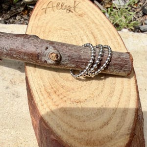Puede incluir: Tres anillos plateados con cuentas exhibidos en una rama de madera, descansando sobre una rodaja de madera. Los anillos est&aacute;n hechos de peque&ntilde;as cuentas redondas. La palabra "Allyk" est&aacute; escrita a mano en la rodaja de madera.