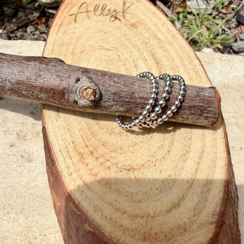 Puede incluir: Tres anillos plateados con cuentas exhibidos en una rama de madera, descansando sobre una rodaja de madera. Los anillos est&aacute;n hechos de peque&ntilde;as cuentas redondas. La palabra "Allyk" est&aacute; escrita a mano en la rodaja de madera.