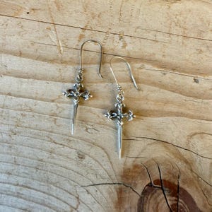 May include: A pair of silver-toned drop earrings featuring a cross-shaped design with ornate detailing and a pointed bottom edge. Each earring is suspended from a curved wire hook, showcasing a metallic finish with intricate, textured metalwork.