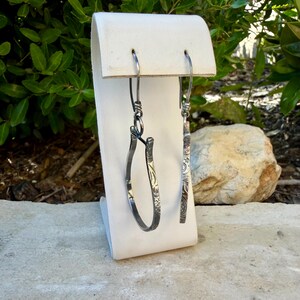 May include: A pair of silver earrings displayed on a white stand. One earring is a curved hoop with an etched floral design. The other is a straight bar with a similar etched pattern. Both earrings have a hook closure.