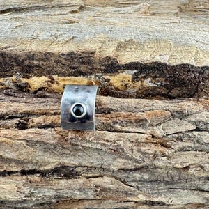 May include: A silver-toned ring with a curved band and a small, round, black stone set in the center. The ring is displayed on a textured, brown wooden surface, creating a rustic contrast.