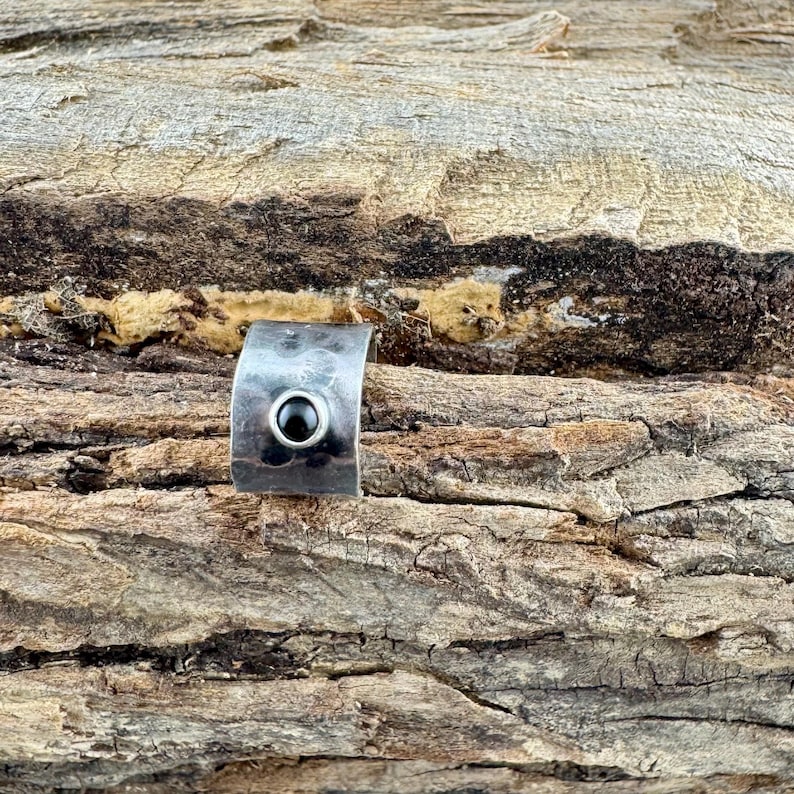 May include: A silver-toned ring with a curved band and a small, round, black stone set in the center. The ring is displayed on a textured, brown wooden surface, creating a rustic contrast.