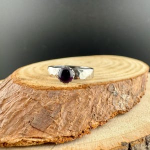 May include: A silver ring with a hammered band and a round, dark purple gemstone. The ring is displayed on a wooden surface against a dark backdrop, highlighting the jewellery's design and colour.