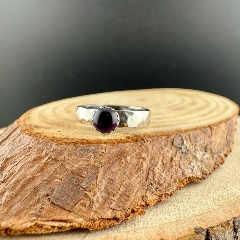 May include: A silver ring with a hammered band and a round, dark purple gemstone. The ring is displayed on a wooden surface against a dark backdrop, highlighting the jewellery's design and colour.