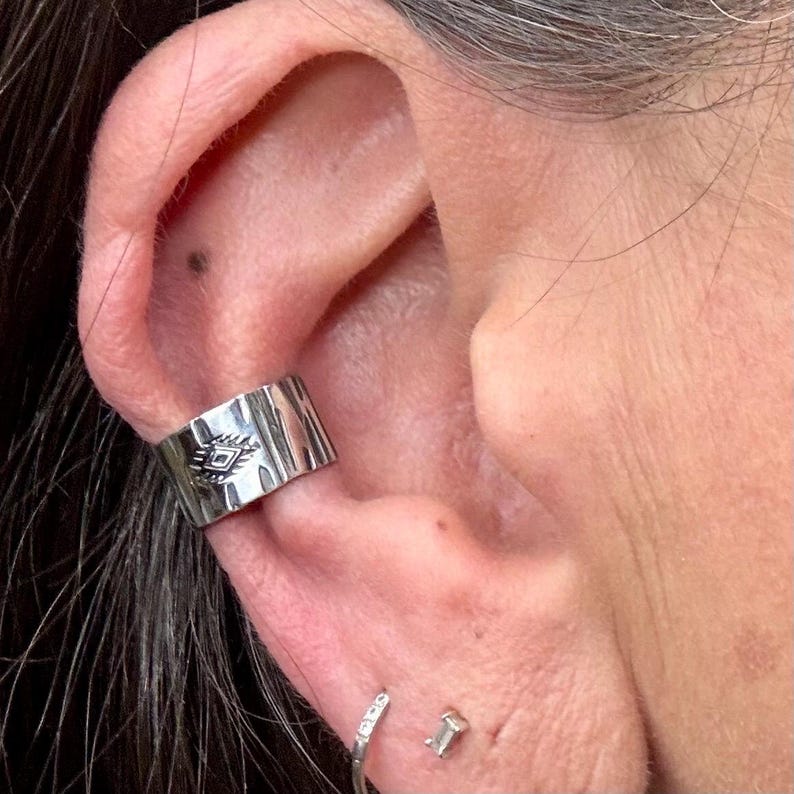 May include: Silver ear cuff with a tribal design. The cuff is worn on the ear and features a geometric pattern. A small, silver earring with a clear stone is also visible.