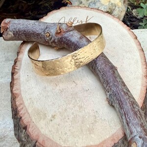 Puede incluir: Un brazalete tipo pu&ntilde;o de metal dorado con una superficie martillada y texturizada. El brazalete es abierto y presenta un acabado pulido que refleja la luz sobre su exterior irregular y con hoyuelos. La pieza se muestra sobre una rama de madera.