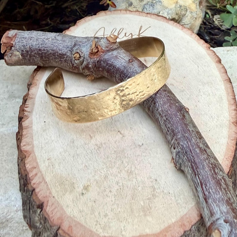 Puede incluir: Un brazalete tipo pu&ntilde;o de metal dorado con una superficie martillada y texturizada. El brazalete es abierto y presenta un acabado pulido que refleja la luz sobre su exterior irregular y con hoyuelos. La pieza se muestra sobre una rama de madera.