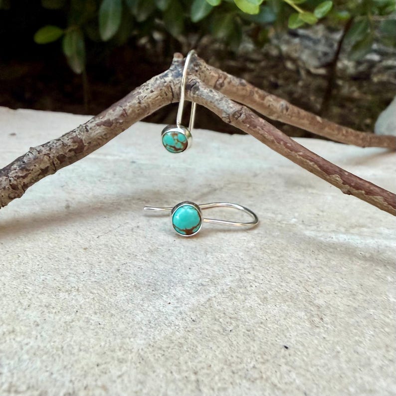 May include: A pair of silver drop earrings featuring round turquoise stones with brown matrix patterns. Each earring consists of a polished silver wire hook that holds a single circular turquoise cabochon, showcasing a bright blue-green color.