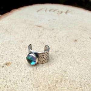 May include: A silver-toned adjustable ring with a round, iridescent gemstone. The ring band features an etched floral design. The gemstone reflects shades of blue, green, and purple. The ring is displayed on a wooden surface.
