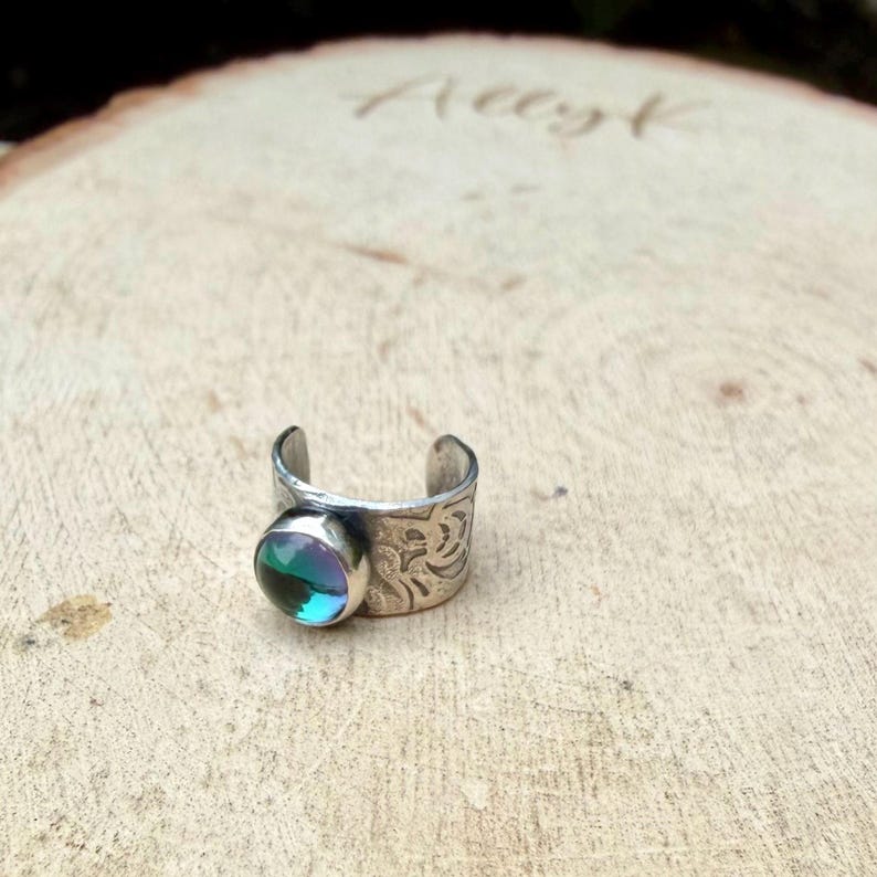 May include: A silver-toned adjustable ring with a round, iridescent gemstone. The ring band features an etched floral design. The gemstone reflects shades of blue, green, and purple. The ring is displayed on a wooden surface.