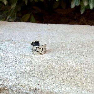 May include: A silver-toned metal ring with an open band design featuring a hammered texture and a stamped outline of the state of Texas on the front surface. The ring has a polished finish and a wide, adjustable band structure.