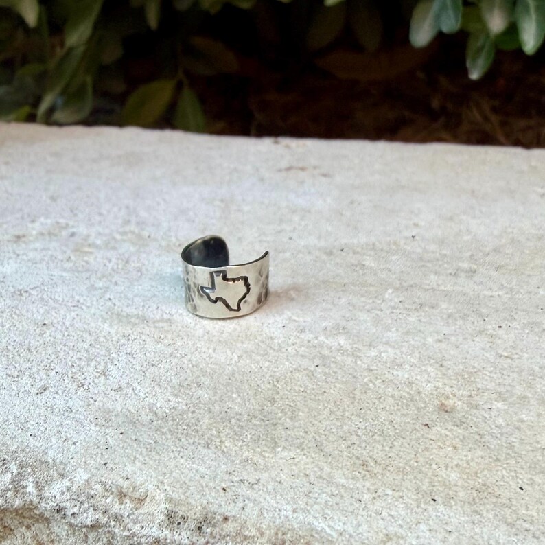 May include: A silver-toned metal ring with an open band design featuring a hammered texture and a stamped outline of the state of Texas on the front surface. The ring has a polished finish and a wide, adjustable band structure.