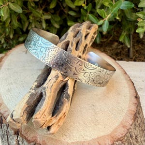 May include: A silver-toned metal cuff bracelet featuring an intricate, engraved pattern of swirling lines, small stars, and geometric shapes. The bracelet has a wide, open-ended band design with a polished finish that reflects light across its surface.