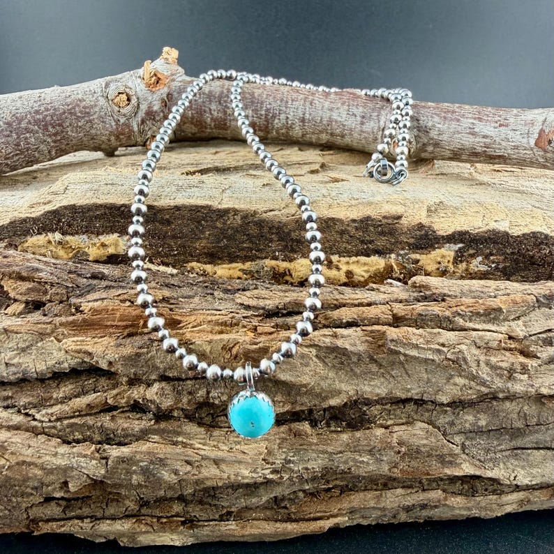 May include: A silver beaded necklace with a turquoise pendant. The necklace is composed of small, round silver beads and features a bright blue, spherical turquoise stone as a focal point. The necklace is displayed on a piece of wood.