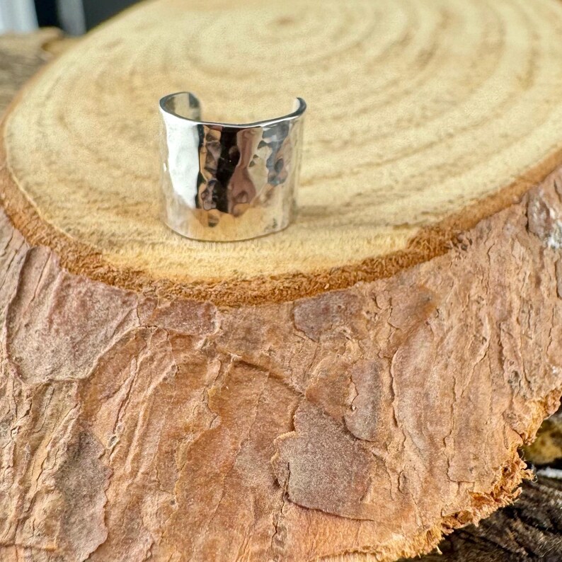 May include: A silver-colored ring with a hammered texture. The ring has a wide band and a curved top, sitting on a wooden surface. The ring's surface reflects light, creating highlights and shadows.
