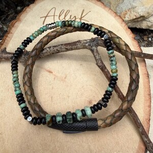 May include: A handcrafted bracelet featuring a braided brown leather band and a strand of black beads interspersed with turquoise-colored stones. The bracelet has a black magnetic clasp and the word "Allyk" is visible in the background.