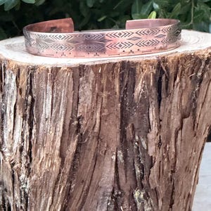 May include: A copper cuff bracelet featuring a repeating geometric pattern of etched diamonds, chevrons, and horizontal lines. The metal has a polished finish with darkened recessed areas that highlight the intricate stamped design details.