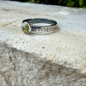 May include: A silver ring with a round, light green and brown stone. The ring band has etched designs. The stone is set in a scalloped bezel. The ring is displayed on a textured, light-colored surface.