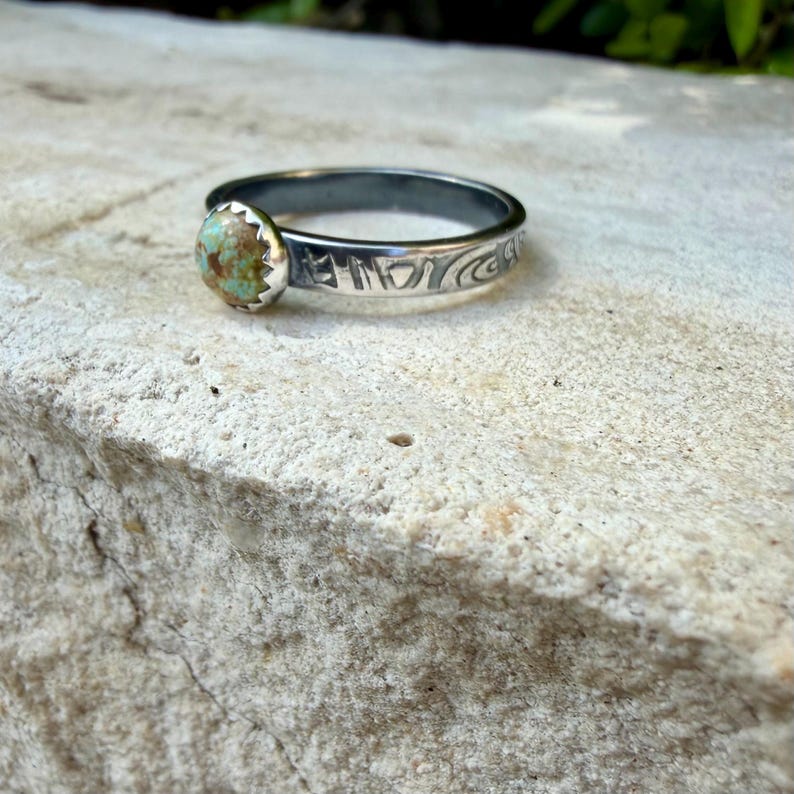 May include: A silver ring with a round, light green and brown stone. The ring band has etched designs. The stone is set in a scalloped bezel. The ring is displayed on a textured, light-colored surface.