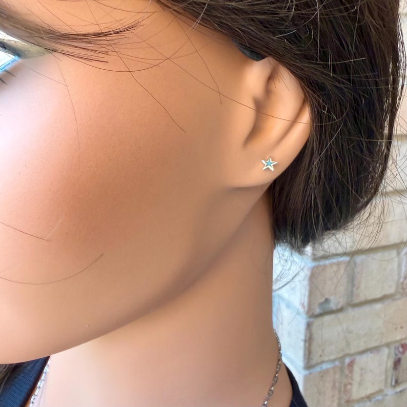 May include: A close-up of a person's ear with a small star-shaped earring. The earring is silver-colored with a light blue center. The person is wearing a dark-colored top and a silver necklace.