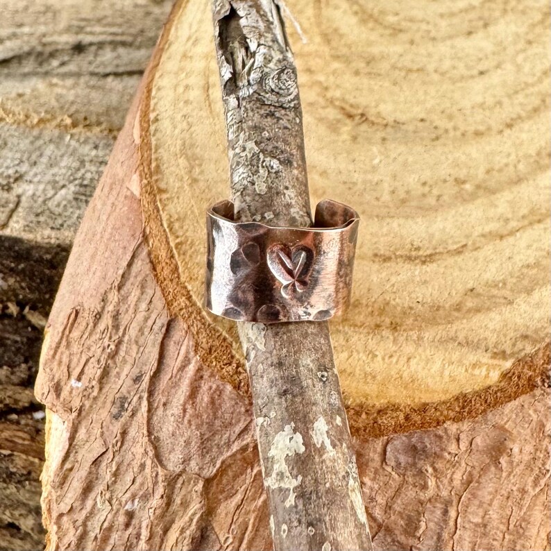May include: A hammered copper ring with a heart-shaped design. The ring is wide and has a textured surface. The heart is stamped into the metal. The ring is displayed on a wooden branch.