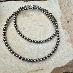 May include: A silver beaded necklace with a double strand design. The necklace features small, round beads and a silver clasp. The jewelry is displayed on a light-colored, textured surface.