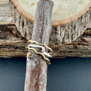 Puede incluir: Tres anillos, dos dorados y uno plateado, se exhiben en un palo de madera. Los anillos dorados tienen un dise&ntilde;o ondulado, mientras que el anillo plateado tiene una banda recta. Los anillos se colocan sobre un fondo de madera y una superficie oscura.