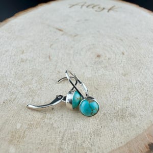 May include: Silver leverback earrings featuring round turquoise stones. The turquoise stones display a mottled pattern of blue and brown. The earrings are presented on a light-coloured wooden surface. The word "Allyk" is etched into the wood.
