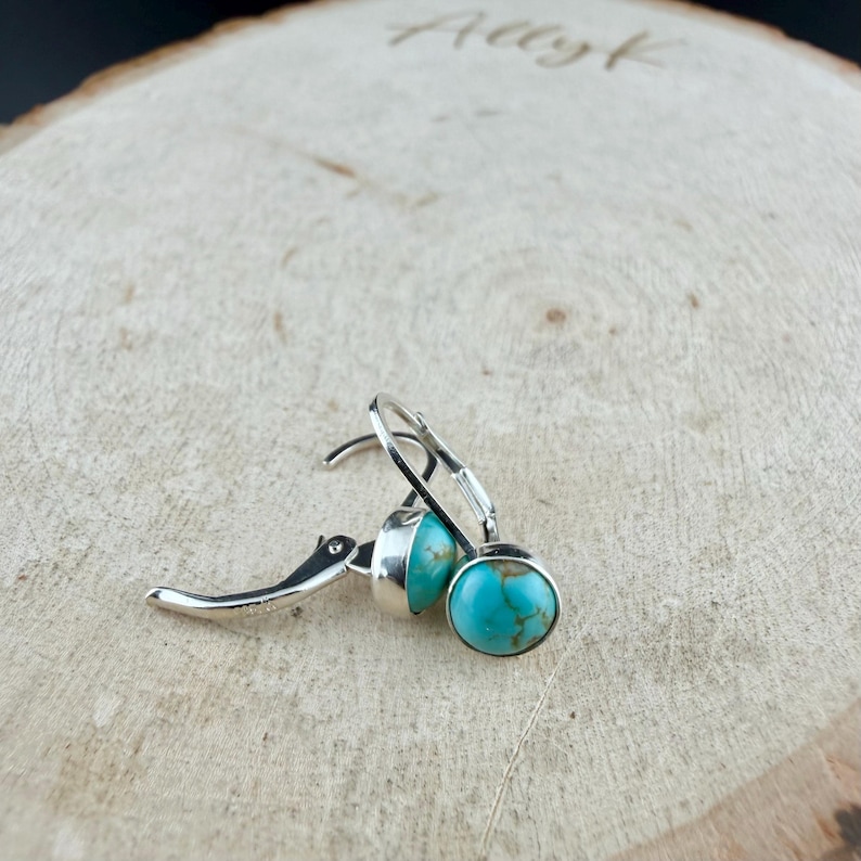 May include: Silver leverback earrings featuring round turquoise stones. The turquoise stones display a mottled pattern of blue and brown. The earrings are presented on a light-coloured wooden surface. The word "Allyk" is etched into the wood.