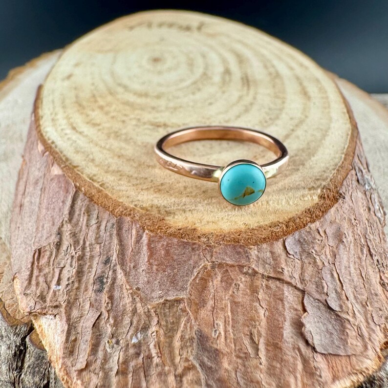 May include: A copper-coloured ring featuring a round, turquoise stone. The ring is set on a wooden surface with a natural, textured appearance. The ring's band has a subtle, textured design.