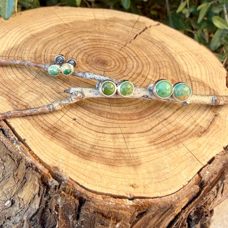 May include: Three pairs of round stud earrings featuring smooth, polished green stones set in silver-toned metal bezels. Each pair displays unique natural variations in the green hues, ranging from light mint to deep forest tones, arranged on a branch.