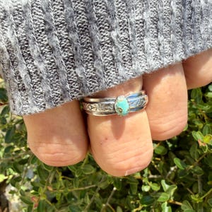 Puede incluir: Un anillo de plata con una peque&ntilde;a piedra turquesa de forma ovalada con inclusiones de matriz marr&oacute;n. La banda est&aacute; hecha de plata pulida y muestra intrincados patrones geom&eacute;tricos grabados a los lados del engaste central de la piedra.