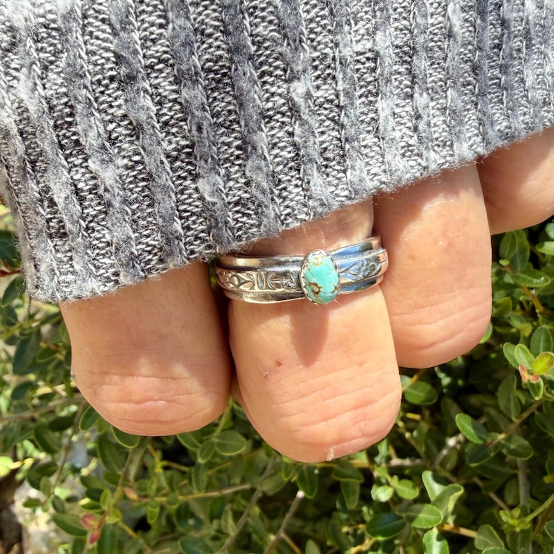 Puede incluir: Un anillo de plata con una peque&ntilde;a piedra turquesa de forma ovalada con inclusiones de matriz marr&oacute;n. La banda est&aacute; hecha de plata pulida y muestra intrincados patrones geom&eacute;tricos grabados a los lados del engaste central de la piedra.