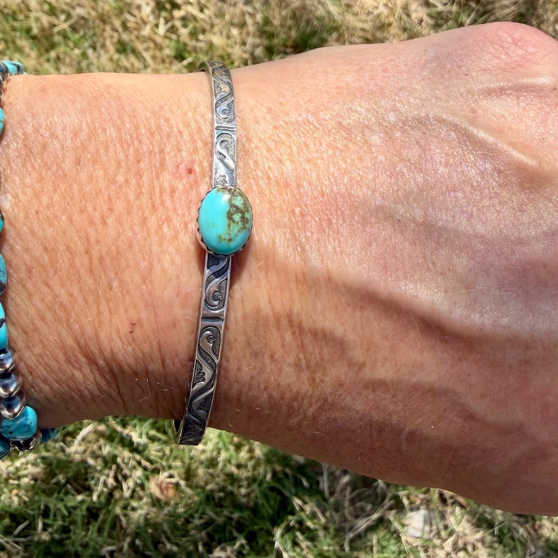 Op de afbeelding: Een zilverkleurige armband met een ovale turkoois steen. De armband heeft een gegraveerd ontwerp langs de zijkanten. Een turkoois kralenarmband is zichtbaar aan de linkerkant van de afbeelding.