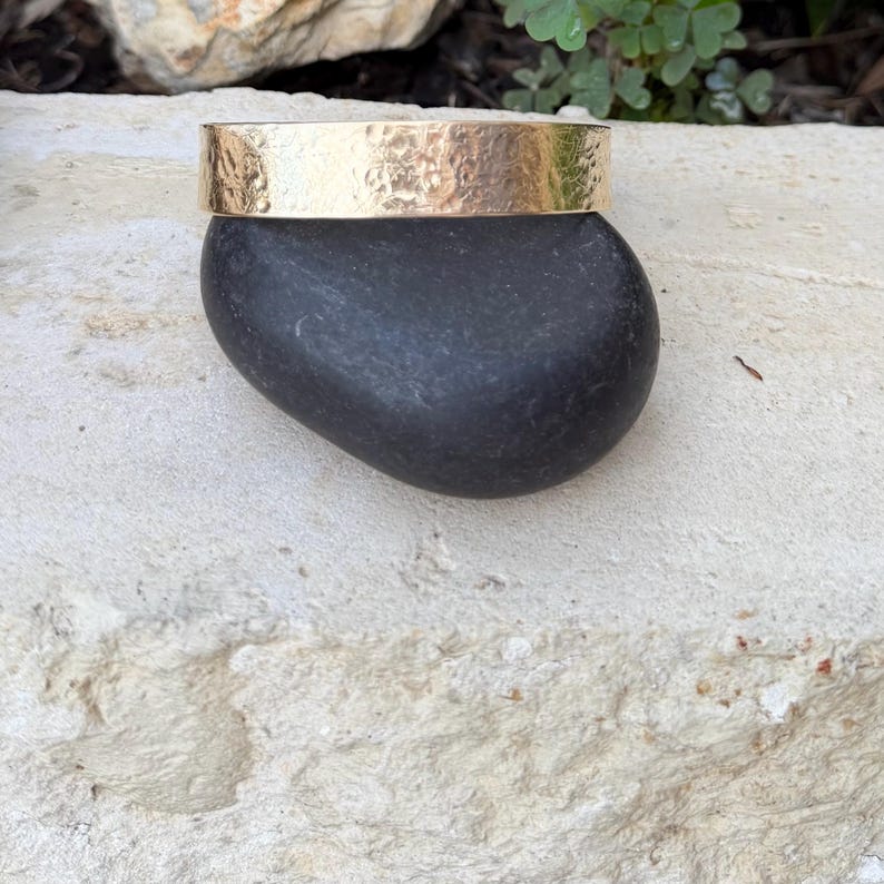 Puede incluir: Una pulsera tipo brazalete de metal dorado con una superficie texturizada y martillada. La banda tiene un perfil ancho y plano con un ancho constante en toda su longitud, mostrando un brillo met&aacute;lico reflectante en la superficie exterior.