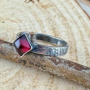 May include: A silver ring with a square, deep red gemstone. The band features etched leaf and geometric patterns. The ring is displayed on a wooden surface, showcasing its handcrafted details and gemstone.