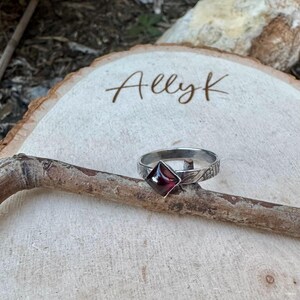 May include: A silver ring with a square, deep red gemstone. The ring band has a textured design. The ring is displayed on a wooden branch, with the name "Allyk" etched on a wooden slice in the background.